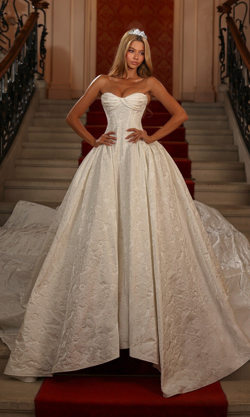 An elegant woman in the ivory Alyce 7164 long white dress stands on a red-carpeted staircase, wearing a tiara and posing with hands on hips.